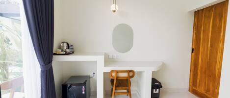 A compact room with a white desk, a wooden chair, a small black refrigerator, a wooden wardrobe, and a hanging light fixture.