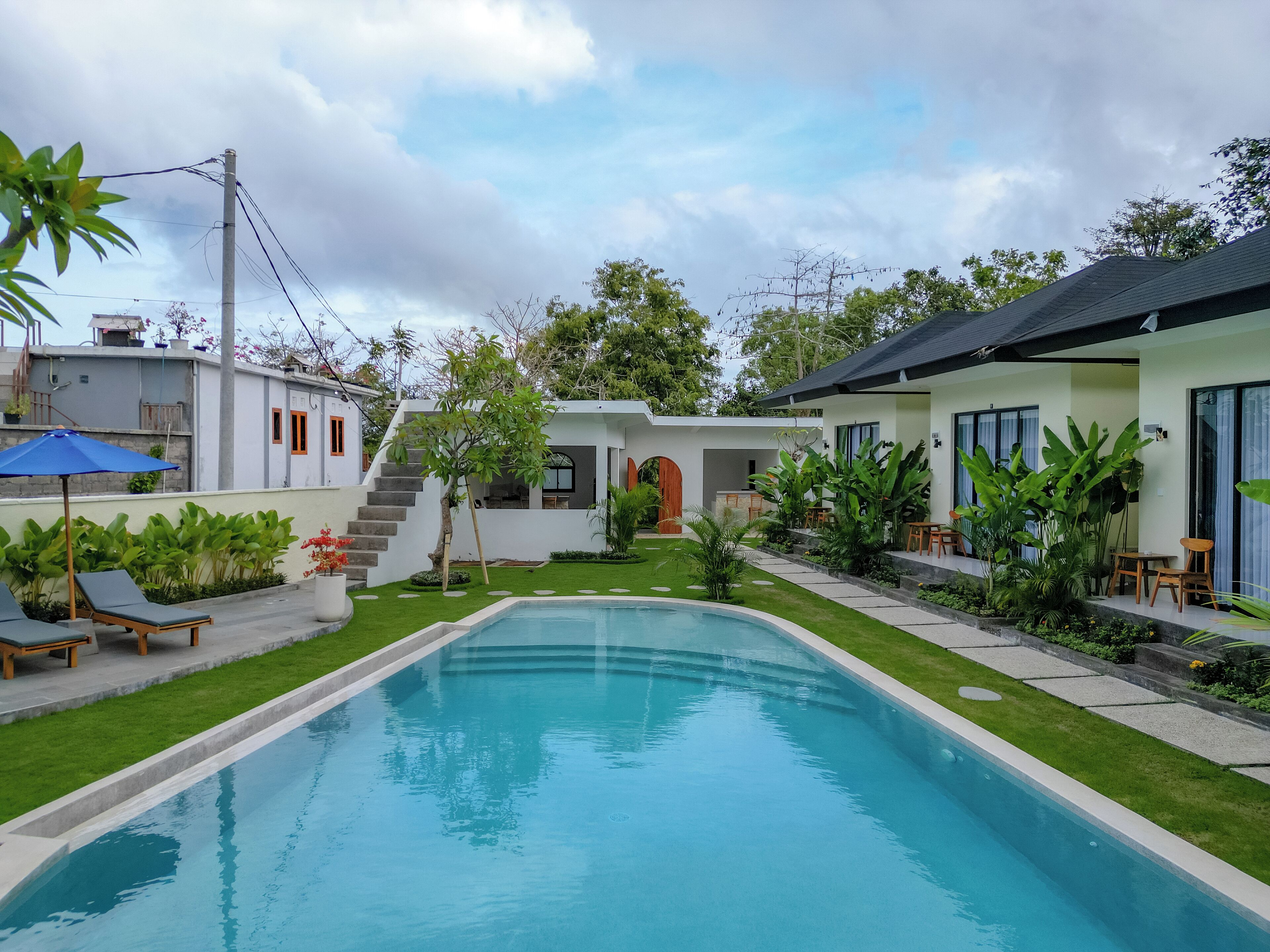 A swimming pool surrounded by a well-maintained lawn, a small garden with various plants, and a modern building with a staircase leading to an upper level.