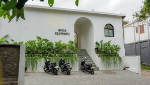 Un bâtiment blanc sur lequel on peut lire l’inscription « KULA ULUWATU ». Trois motos sont garées devant, et un escalier mène à l’entrée.