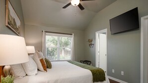 A bedroom with a bed, bedside lamps, a ceiling fan, a window with blinds, a door, and a flat-screen TV mounted on the wall.