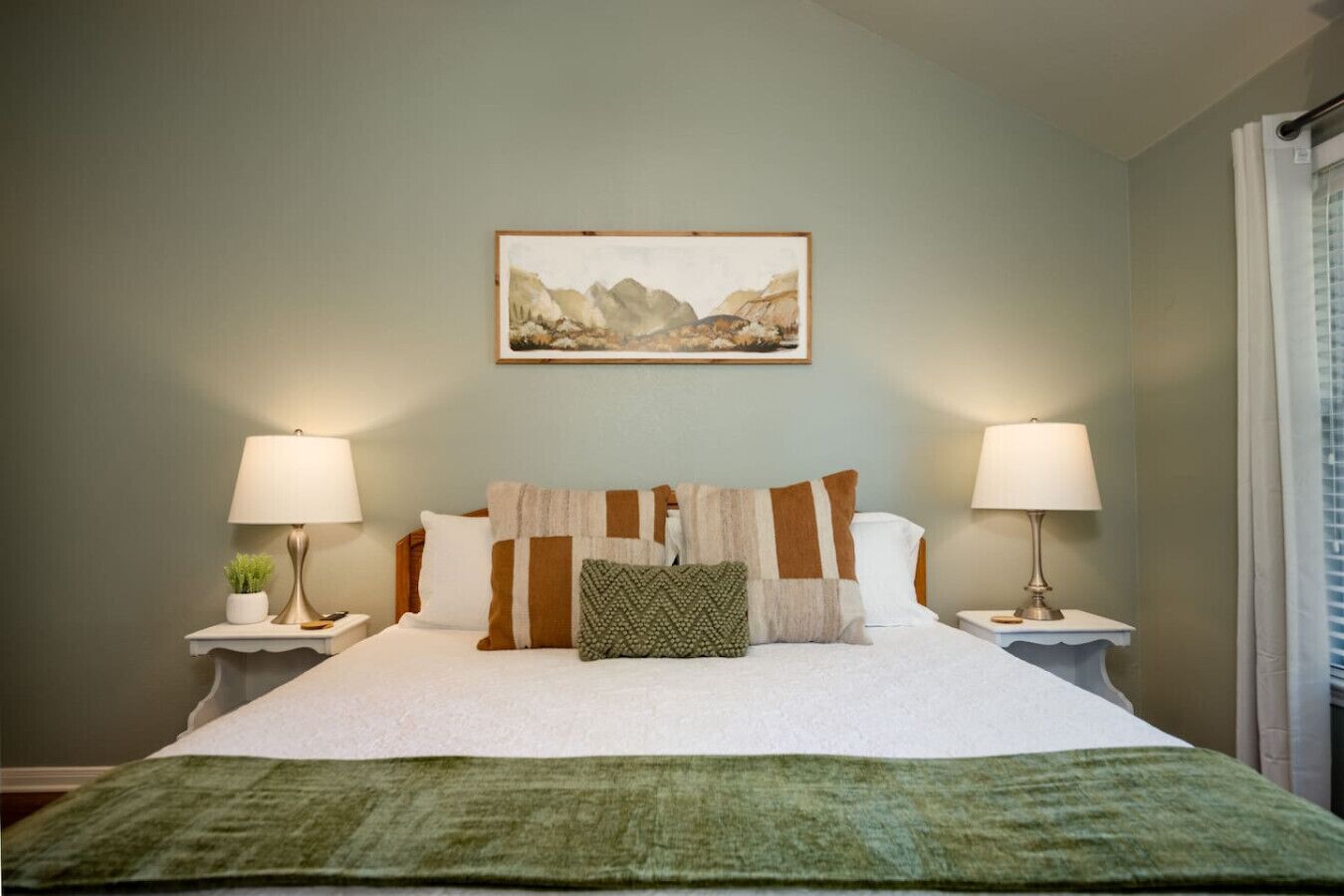 A neatly made bed with a green throw blanket, two bedside lamps, and a framed landscape painting on the wall.