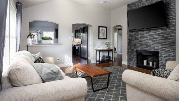 A modern living room with a fireplace, a flat-screen TV mounted on the wall, and a kitchen visible through an archway.