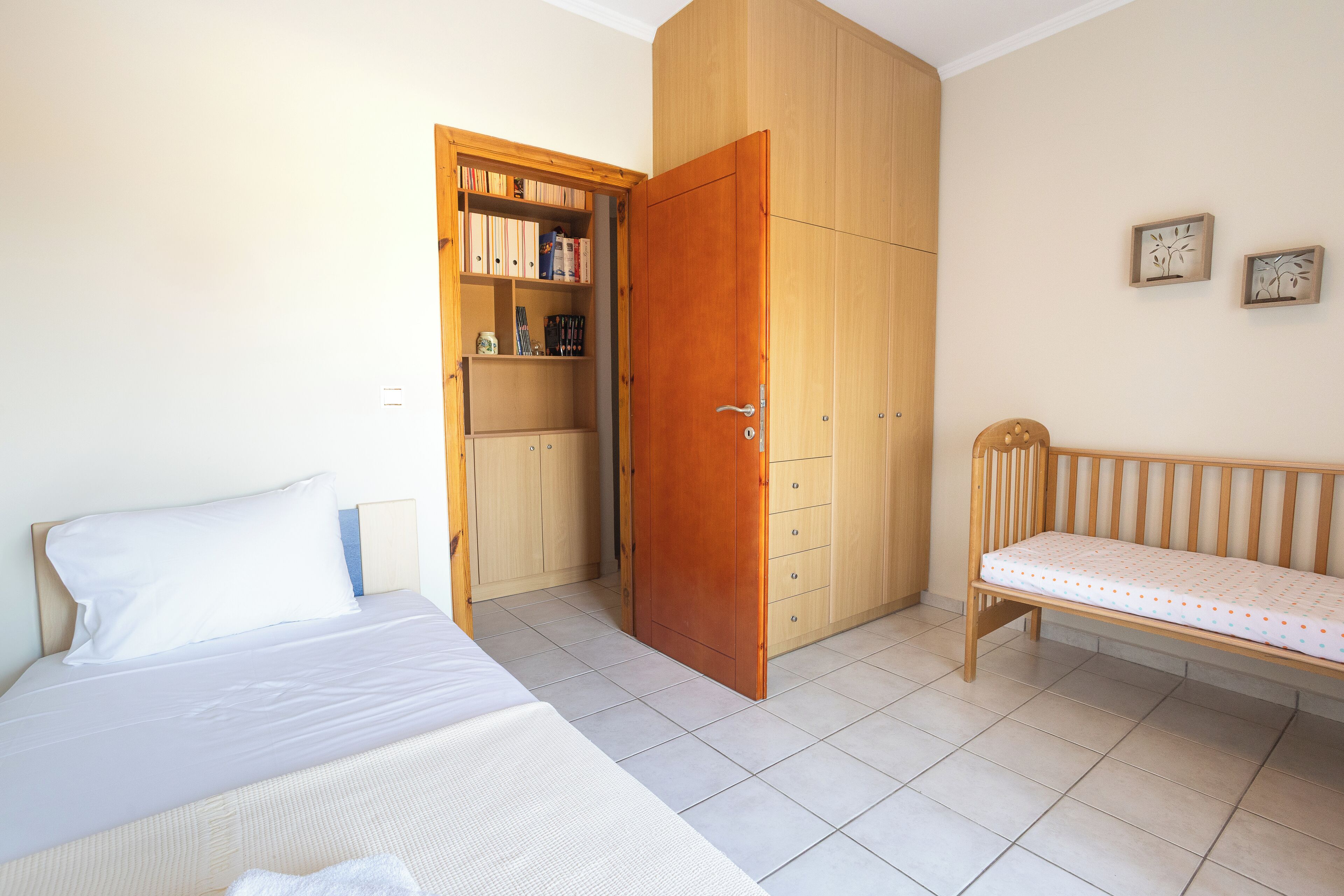 Een slaapkamer met een bed, een houten garderobe, een bankje en een plank met boeken en spullen.