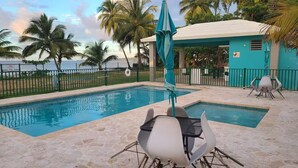 Uma área de piscina com mesa e cadeiras, um espaço coberto para sentar e vista para o mar com palmeiras.