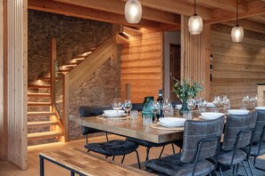 A dining area with a wooden table set for six, surrounded by dark upholstered chairs. A staircase is visible in the background, leading to an upper level. - Chalet OLARICE - Luxe & Spa in Montgenèvre - 12 persons (Montgenèvre)