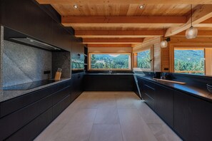 A modern kitchen with dark cabinetry, a granite countertop, and a view of a mountainous landscape through large windows. - Chalet OLARICE - Luxe & Spa in Montgenèvre - 12 persons (Montgenèvre)