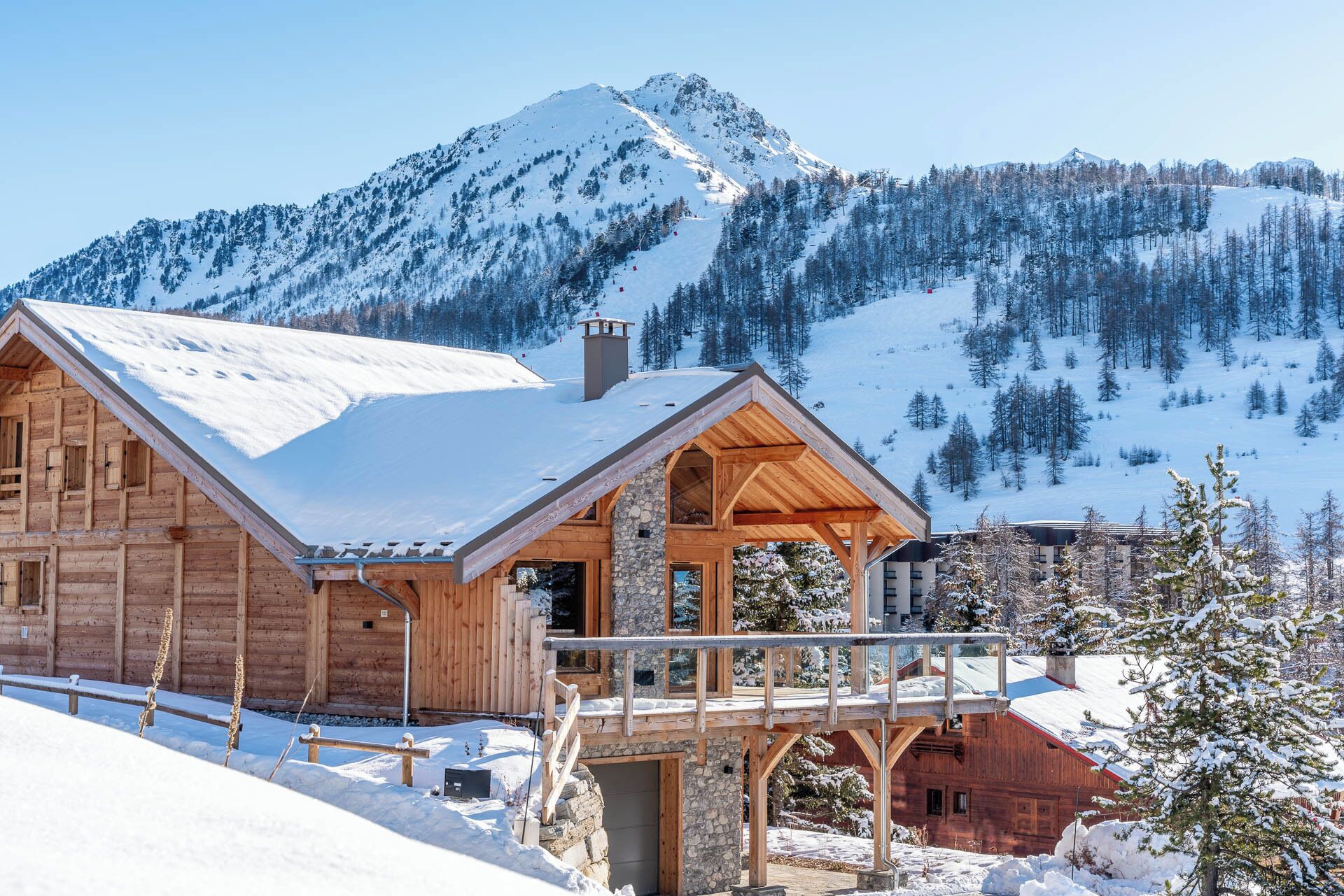 A wooden cabin with a stone foundation, a balcony, and a view of snow-covered mountains.