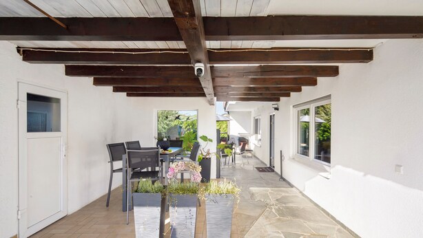 Un porche cubierto al aire libre con suelo de piedra, vigas de madera, una zona de estar con sillas y una mesa, plantas en macetas y vistas de la casa.