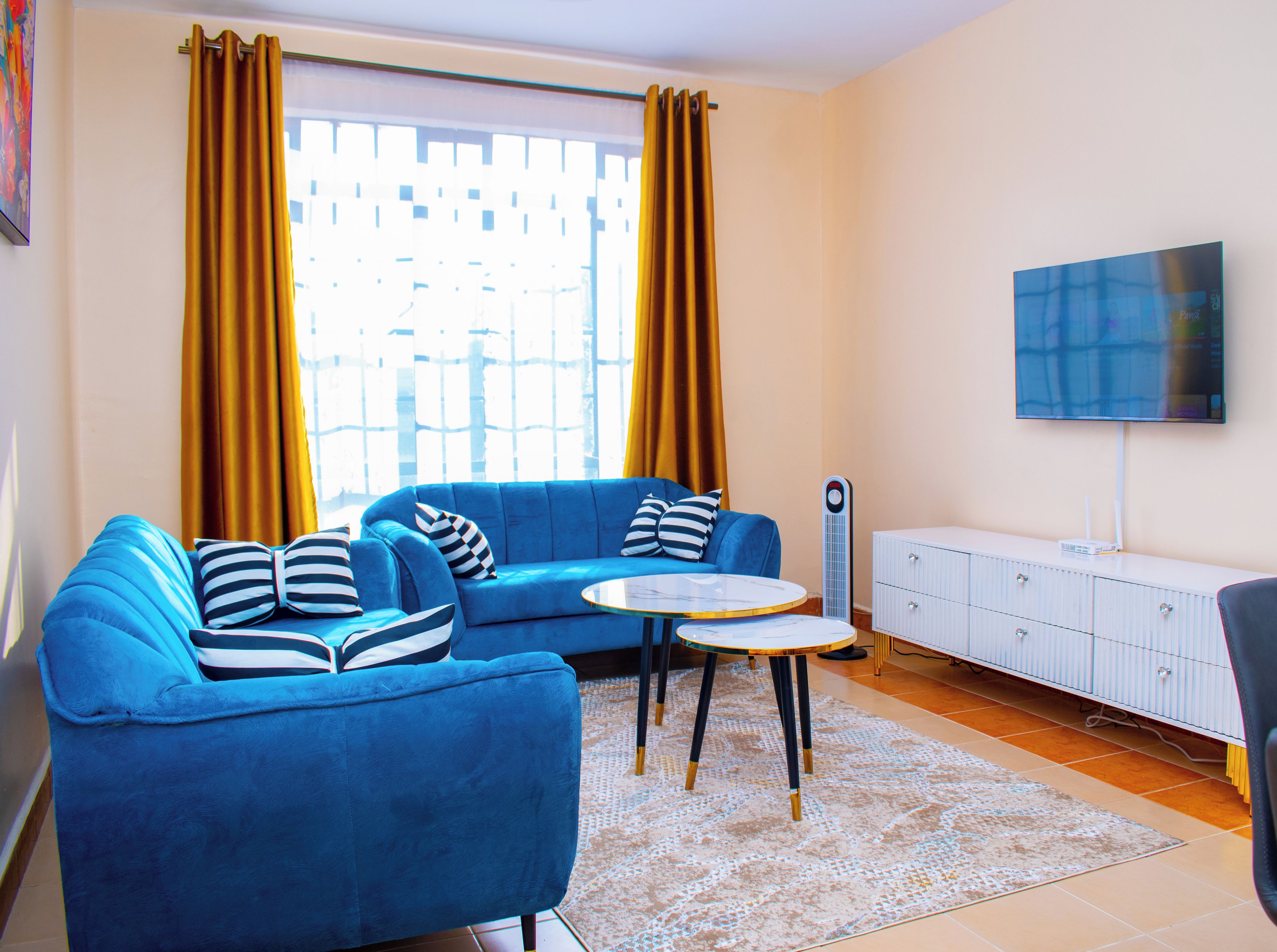 Een woonkamer met een blauwe bank, twee bijzettafels, een wit kastje en een flatscreen televisie aan de muur.