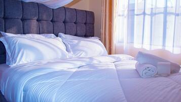 Een net opgemaakt bed met witte lakens en een opgerolde handdoek, een gestofte hoofdbord en een raam met gordijnen.