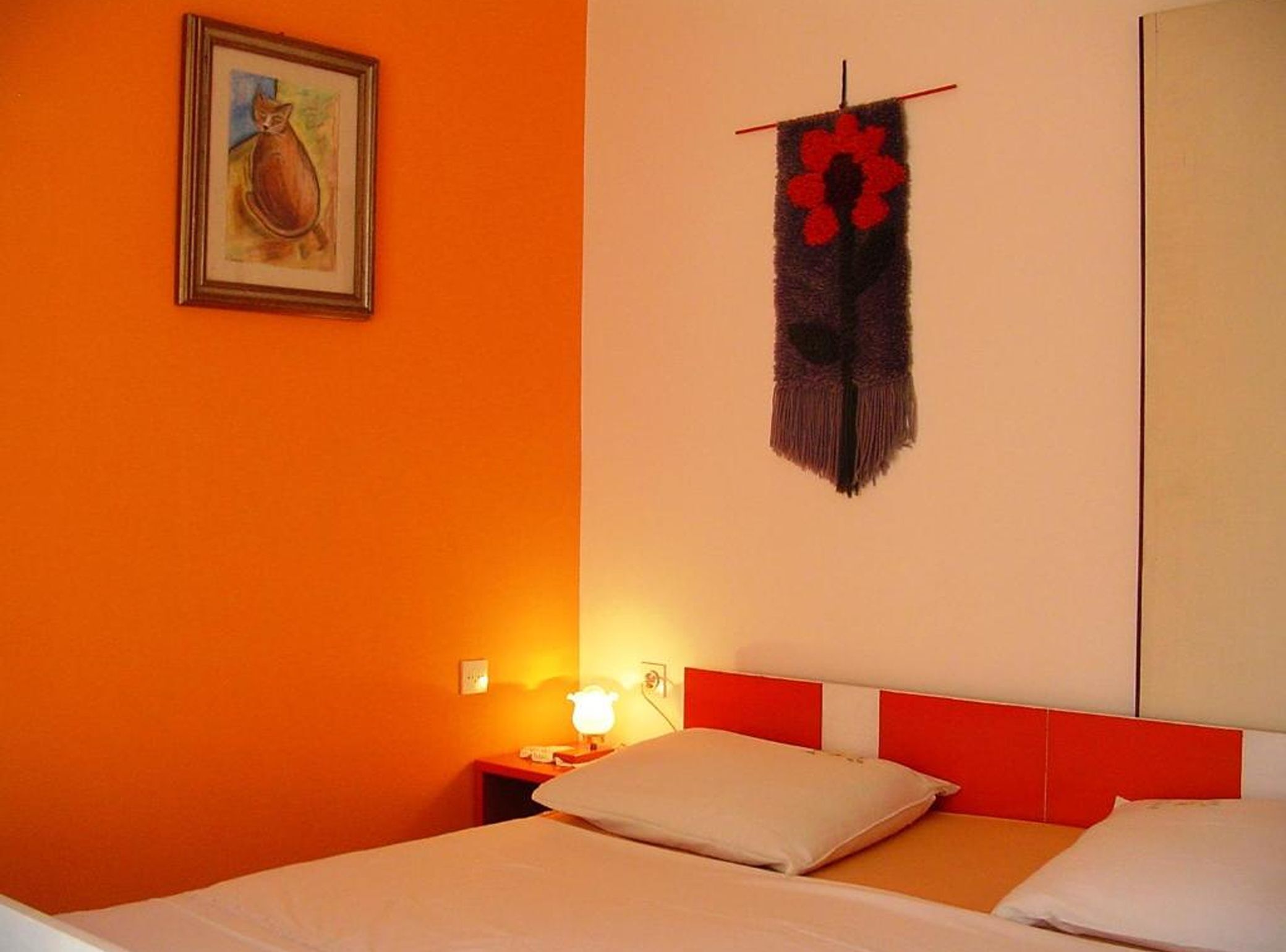 A hotel room with an orange wall, a bed with white bedding, a red headboard, a nightstand with a lamp, and a wall hanging with a red flower.