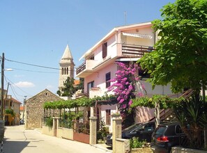 A residential street with a stone building, a pink house with a balcony, and a church tower in the background. - Apartments A&N Pakoštane (Pakostane)