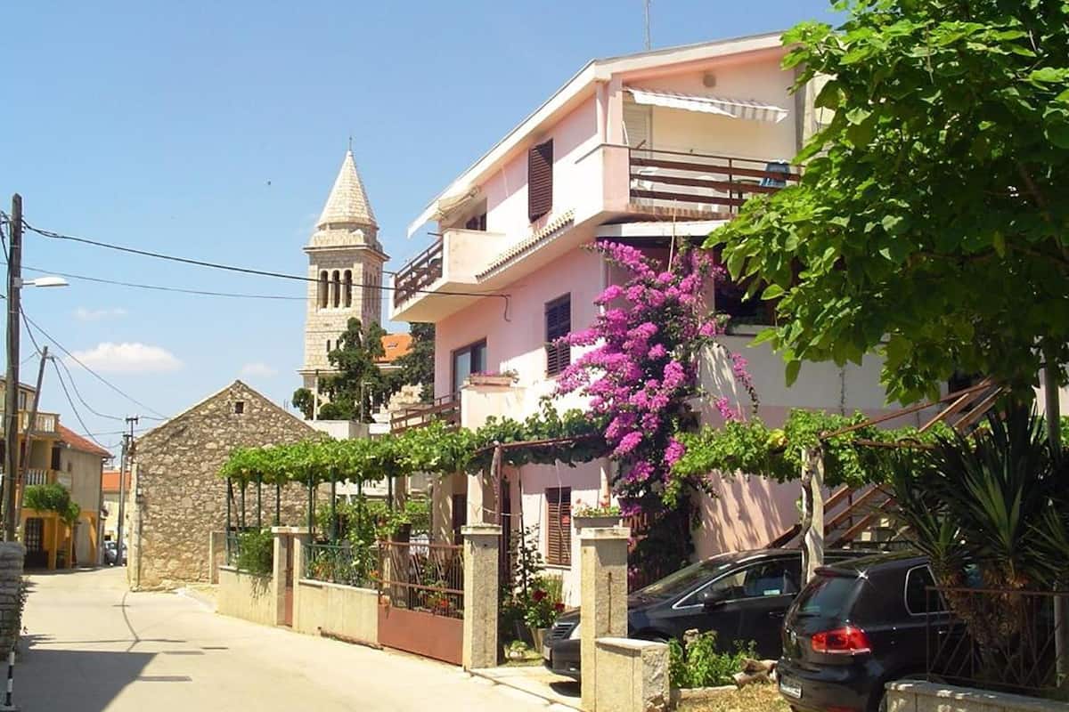 A residential street with a stone building, a pink house with a balcony, and a church tower in the background.