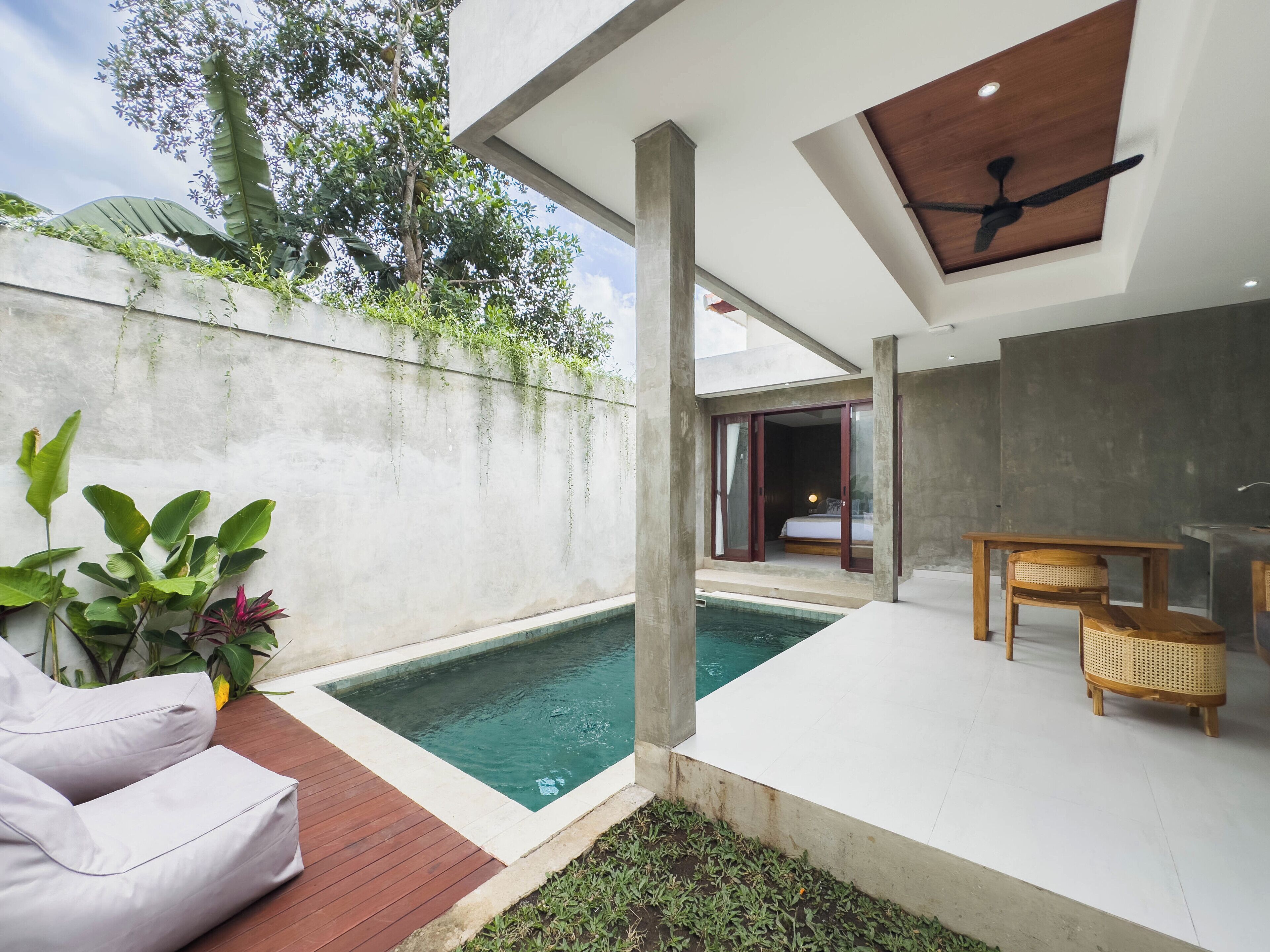 Un área de piscina exterior moderna con un muro de hormigón, una terraza de madera y un ventanal que da a un dormitorio.