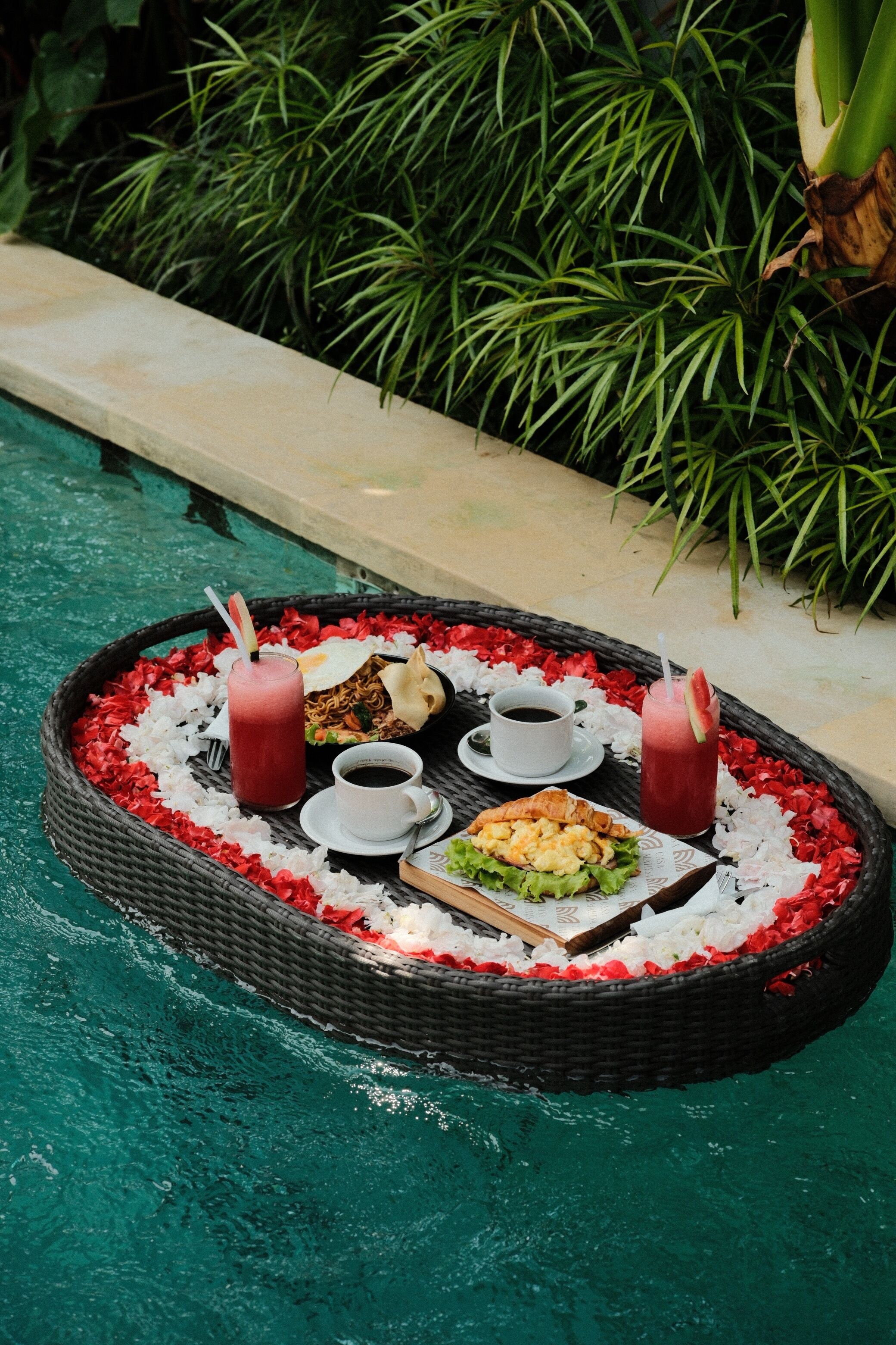 A floating dining setup with a drink, a cup, and a plate of food by a pool.