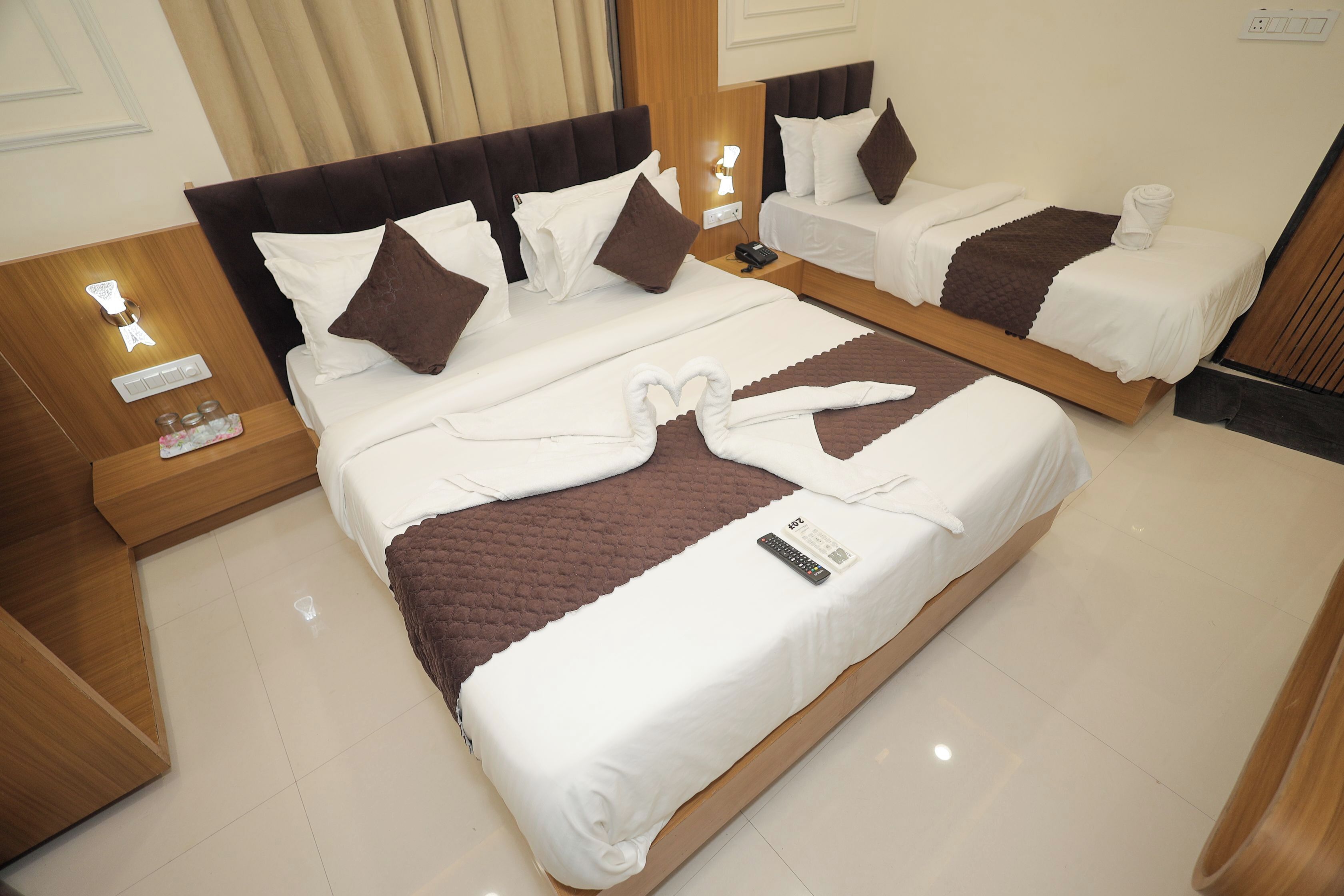 A hotel room with two beds, each with white linens and brown accents. There are bedside tables with lamps and a remote control on the larger bed.
