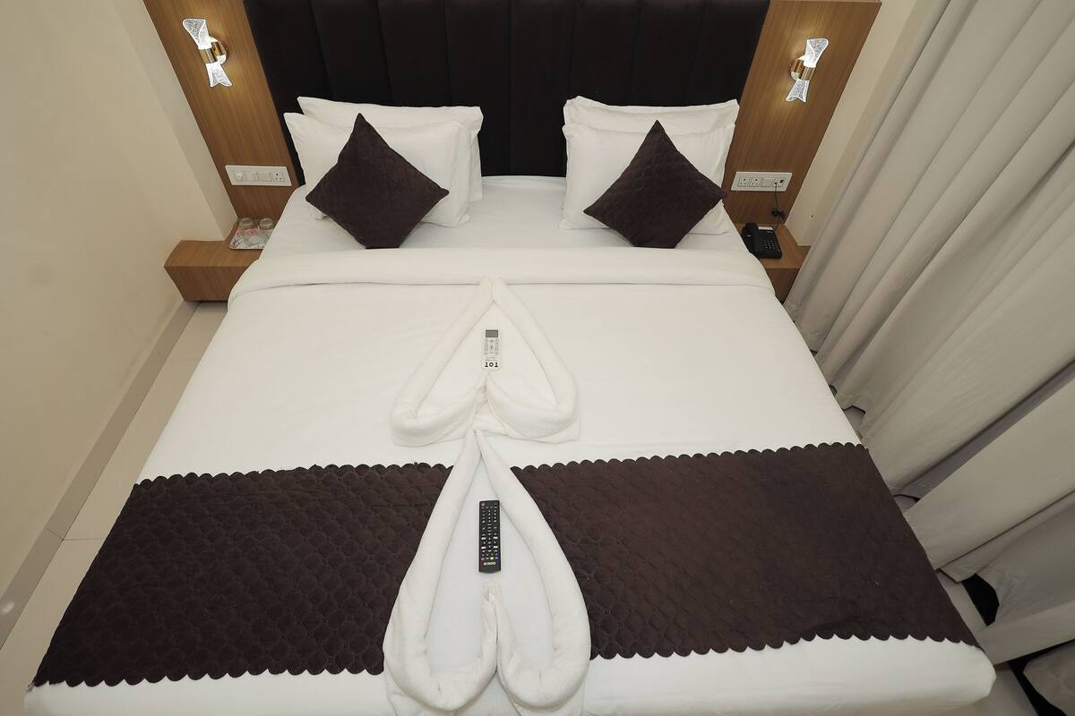 Una cama bien tendida con un control remoto encima, una mesita de noche con un teléfono, y una lámpara de pared.