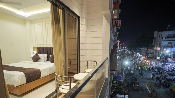 A hotel room with a bed, a chair, and a balcony overlooking a busy street at night. - Hotel Neha Palace (Ujjain)