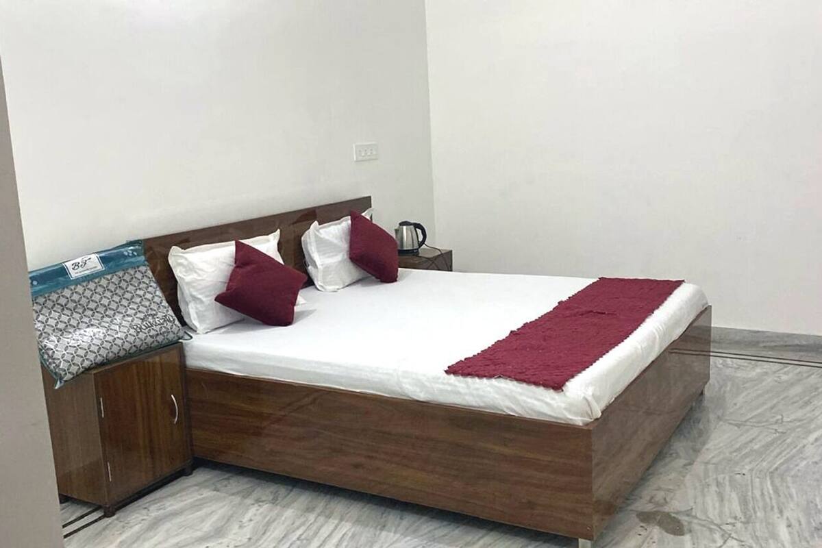 Una cama con estructura de madera, ropa de cama blanca y dos almohadas rojas. Se ve una mesita de noche con una tetera.