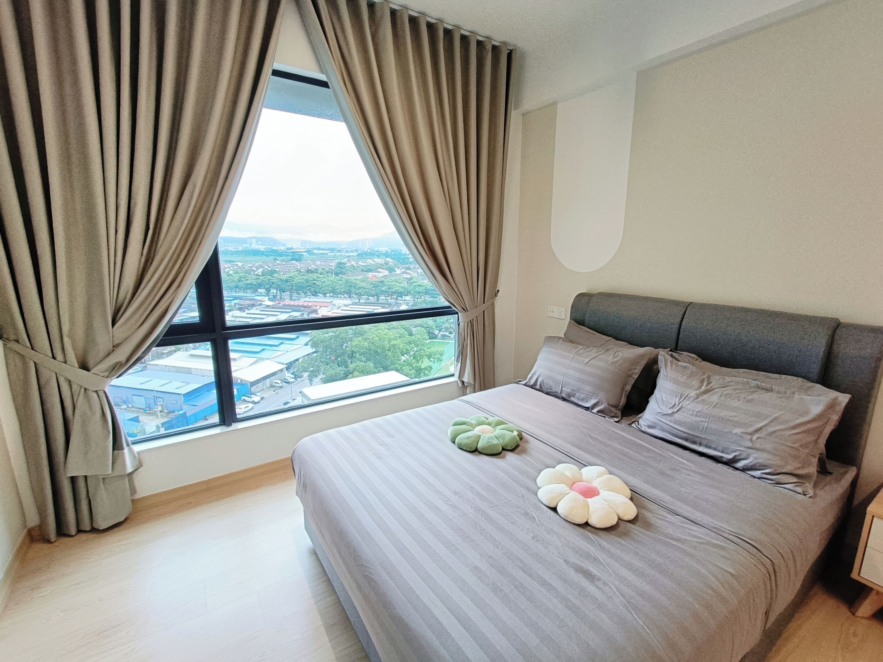 A bedroom with a large window, a bed with grey bedding, and a view of a cityscape.