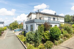 A modern two-story house with a balcony, surrounded by lush greenery and a paved driveway.