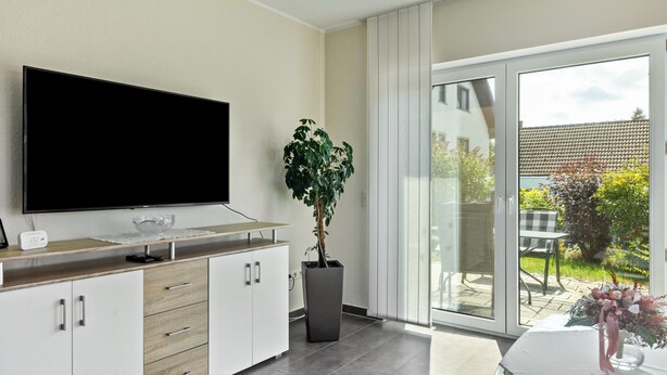 A modern living room with a flat-screen TV, a potted plant, and a sliding glass door leading to an outdoor patio.
