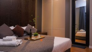 Ein modernes Hotelzimmer mit einem großen Bett, einem Tablett mit Handtüchern und einer Flasche sowie Blick in ein weiteres Zimmer durch eine Glastür.