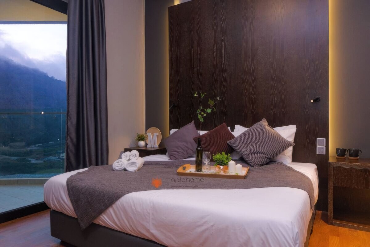 Una habitación de hotel moderna con una cama grande, cabecera de madera, mesita de noche con una bandeja de desayuno y vista a un paisaje montañoso a través de un ventanal amplio.