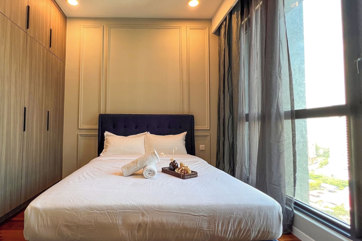 A neatly made bed with white linens and pillows, a dark headboard, and a window with curtains.