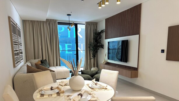 A modern living room with a round dining table, beige sofas, and a flat-screen TV mounted on the wall.