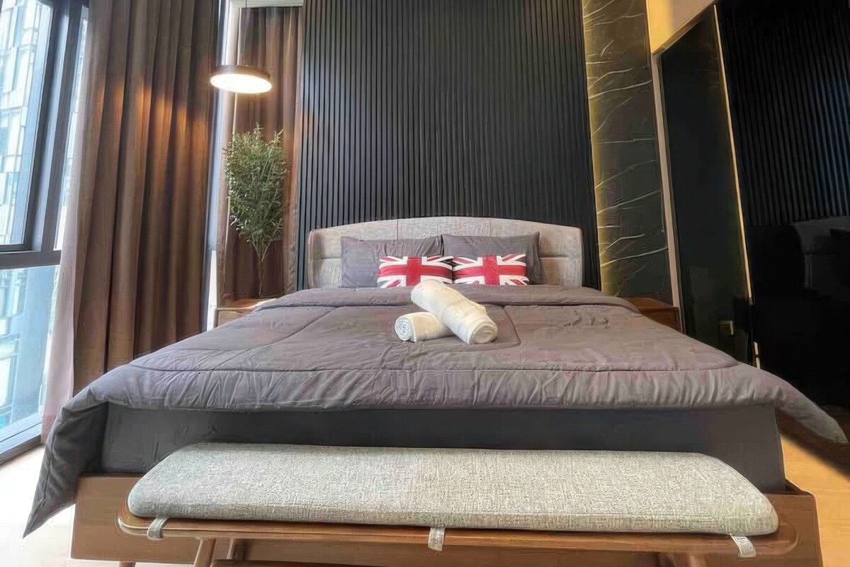 Une chambre moderne avec un grand lit, une table de chevet, une plante en pot et une vue sur la ville grâce à de grandes fenêtres.