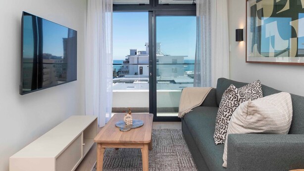 A modern living room with a sofa, a coffee table, a flat-screen TV, and a large window offering a view of buildings and the sea. - The Sage Aparthotel by Totalstay (CAPE TOWN)
