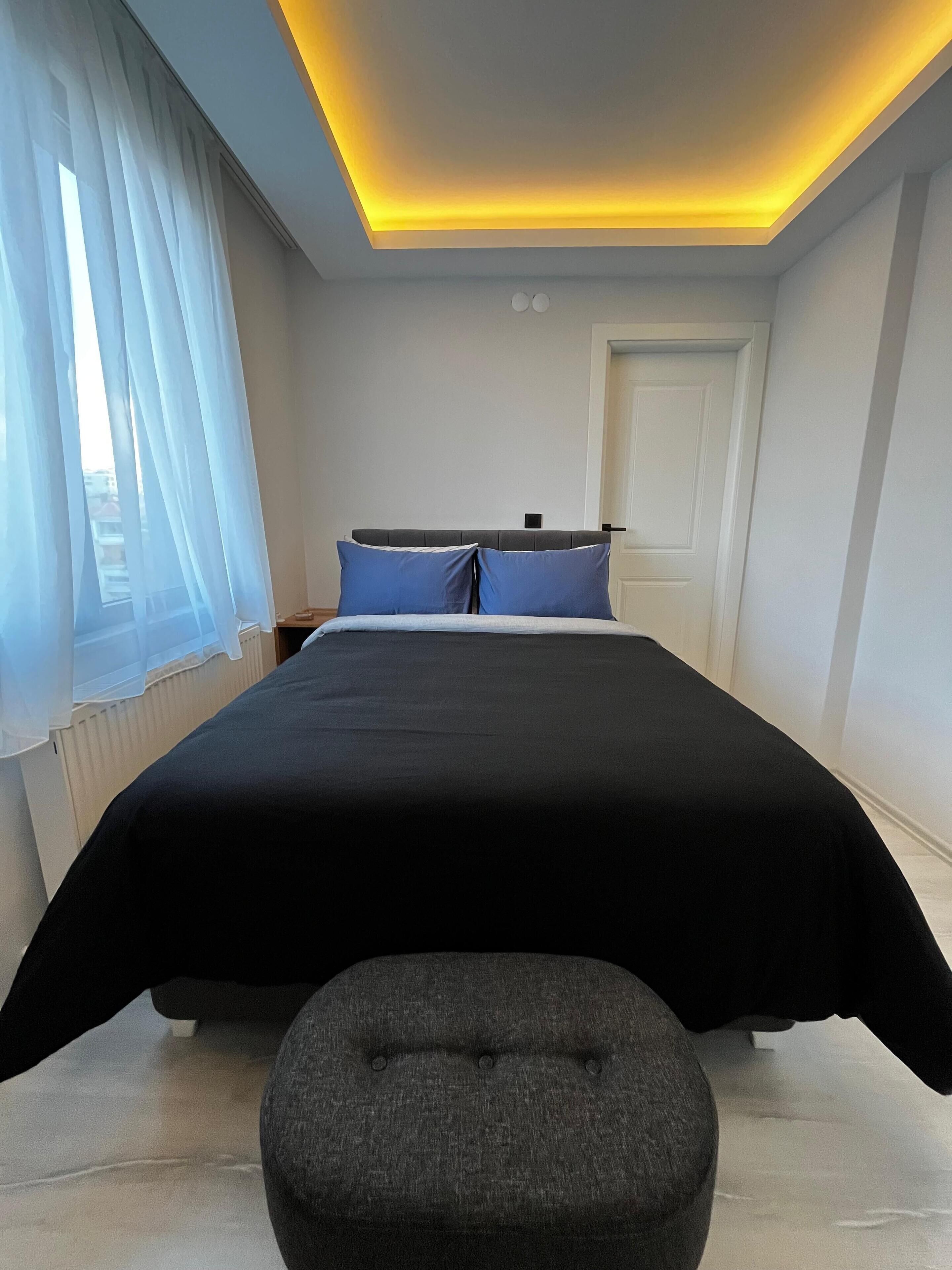 Un dormitorio moderno con una cama, un reposapiés gris e iluminación empotrada en el techo.