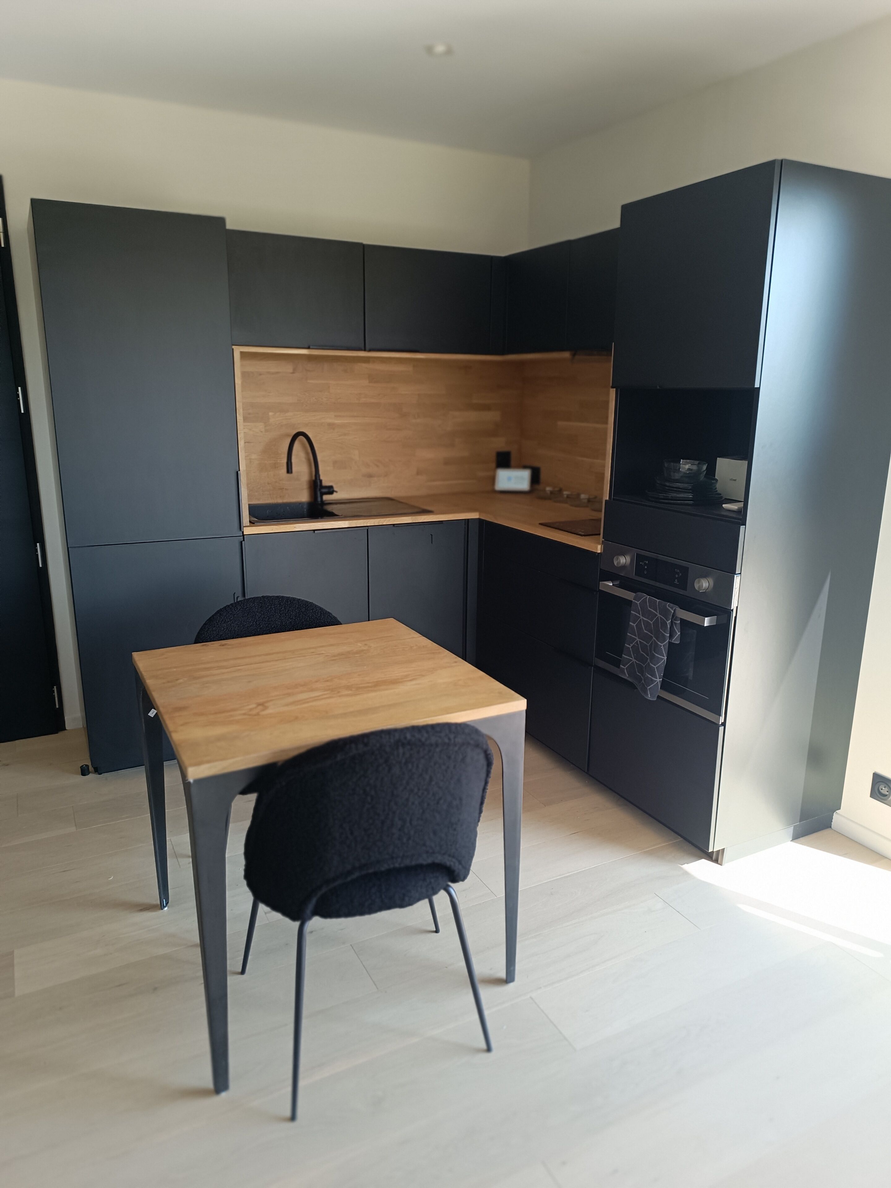 A compact kitchen with a wooden table and black chairs, a built-in oven, and a black cabinet with a wooden shelf.
