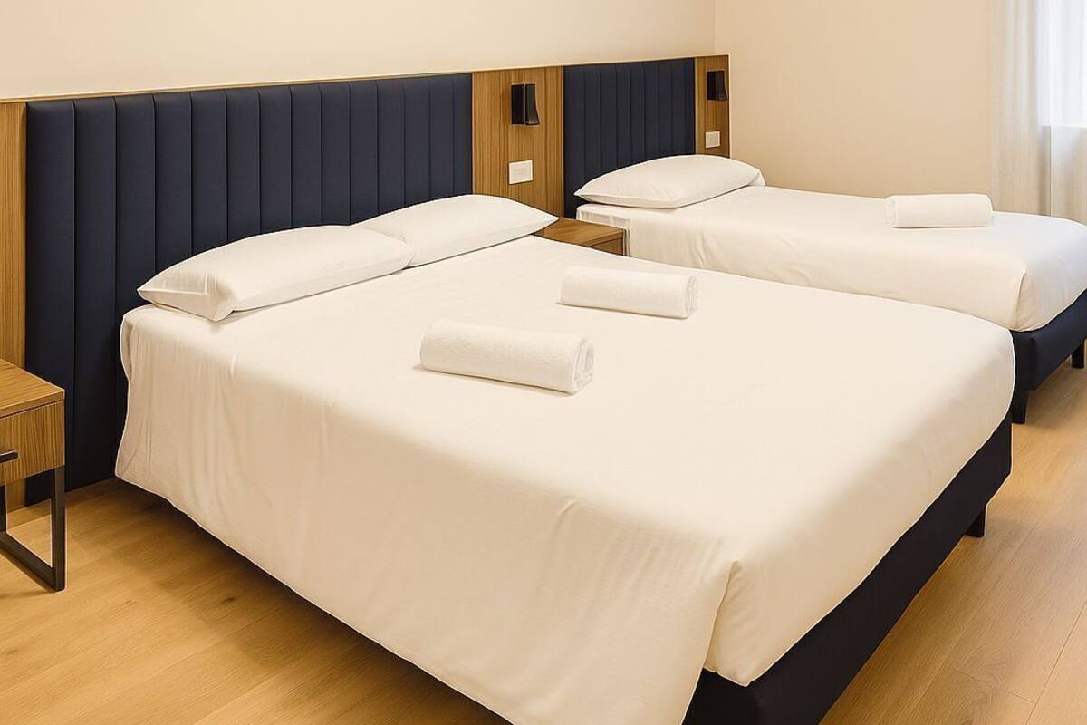 A hotel room with two beds, a wooden nightstand, a modern chandelier, and a window with curtains.