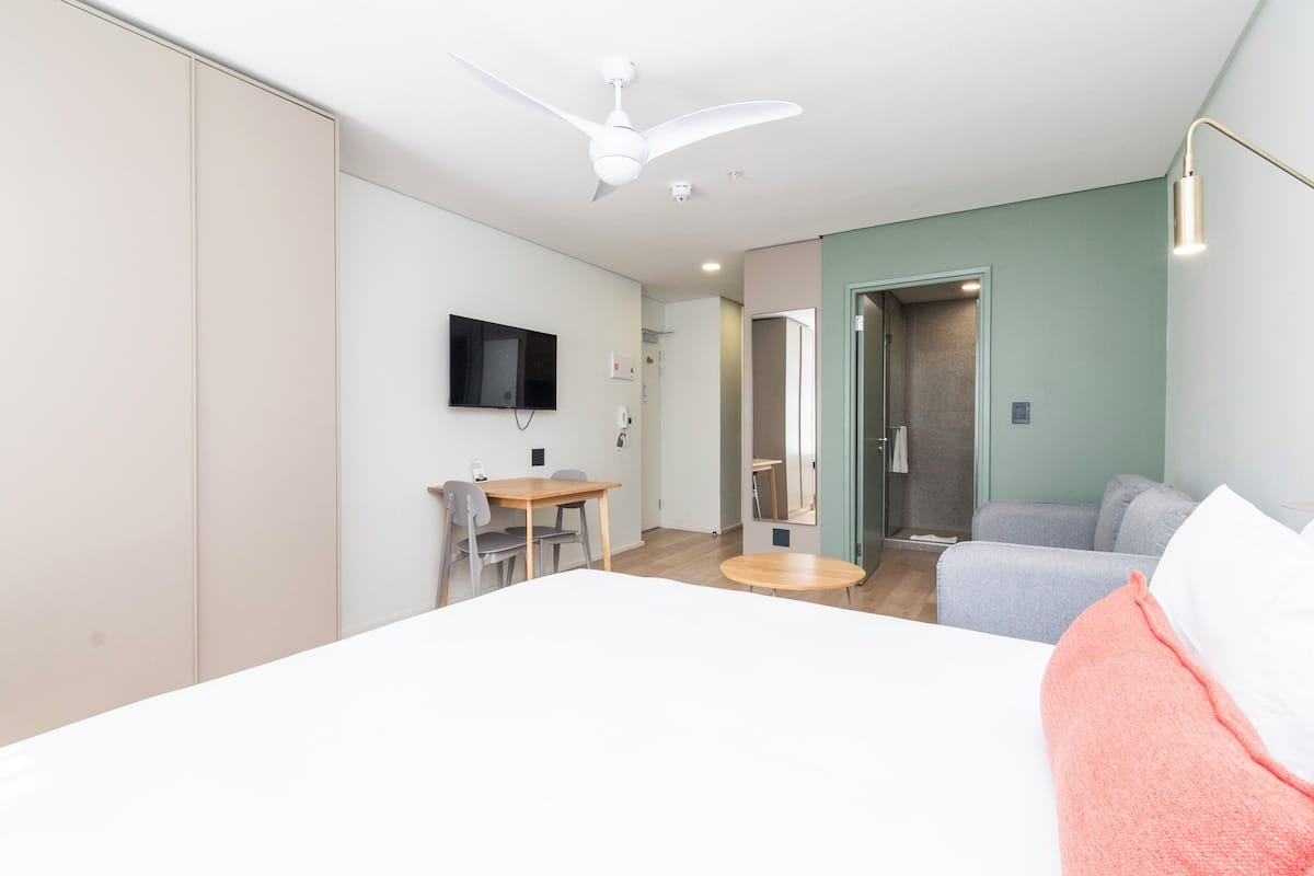 Quarto de hotel moderno com cama, escrivaninha, cadeira, televisão e ventoinha no teto.
