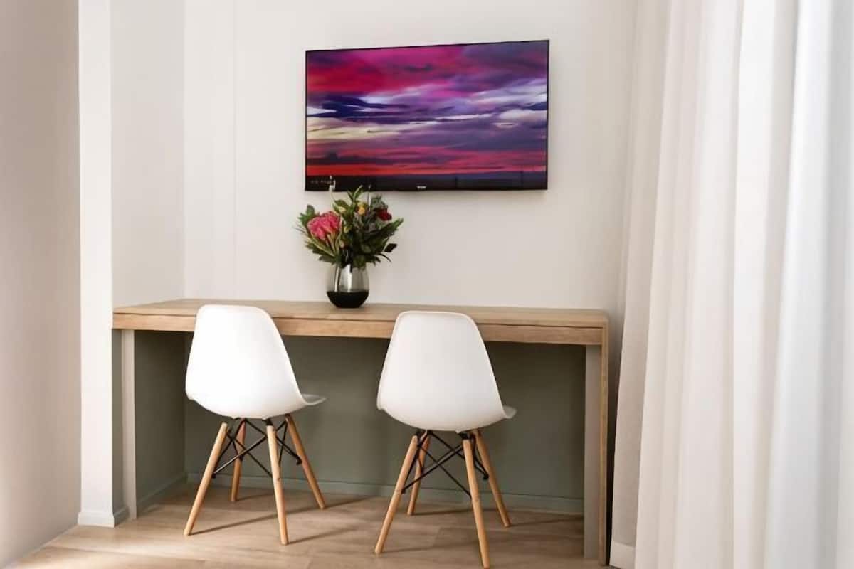 Quarto com escrivaninha de madeira, duas cadeiras brancas, um vaso com flores e uma pintura grande na parede.