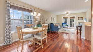 Il y a un coin repas avec une table ronde, des chaises et un lustre. Un coin salon est aménagé avec un canapé et une table basse. La pièce est dotée d’un parquet et de grandes fenêtres.
