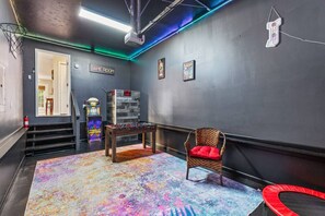 Une pièce comprenant une table de baby-foot, une chaise, un panneau indiquant qu’il s’agit d ’une salle de jeux, un escalier et des jeux d’arcade.