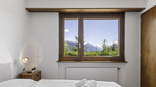 Een slaapkamer met een groot raam, een nachtkastje met een lamp en een netjes opgemaakt bed.