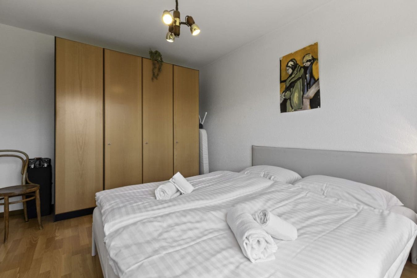 A hotel room with a large bed, a wooden wardrobe, a chair, and a painting on the wall.
