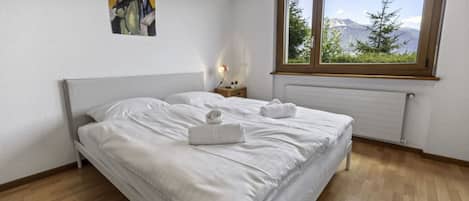 A bedroom with a bed, a nightstand, a window with a view of mountains, and a painting on the wall.