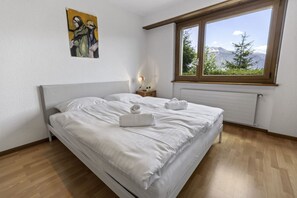 Een slaapkamer met een bed, een nachtkastje, een raam met uitzicht op bergen en een schilderij aan de muur.