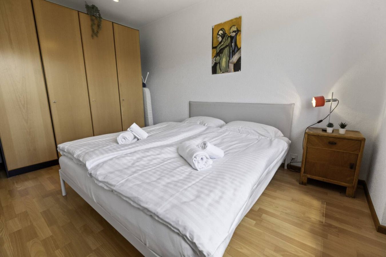 A double bed with white linens and towels, a wooden wardrobe, a bedside table with a lamp, and a small potted plant.