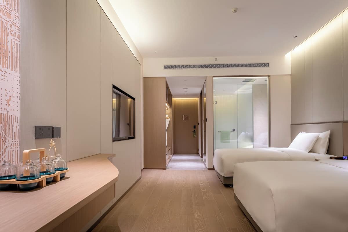 A modern hotel room with a wooden floor, a bed, a bedside table with a lamp, a mirror, and a bathroom with a glass shower enclosure.