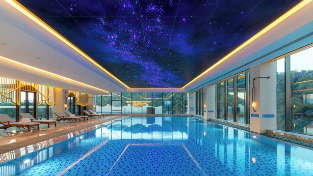 A modern indoor swimming pool with a starry ceiling, sun loungers, and large windows offering a view of the outdoors.