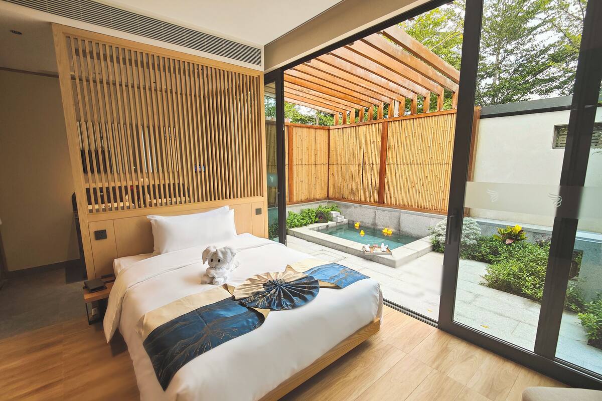 A modern bedroom with a large bed, a small bedside table, a wooden headboard, and a view of a garden and outdoor area.