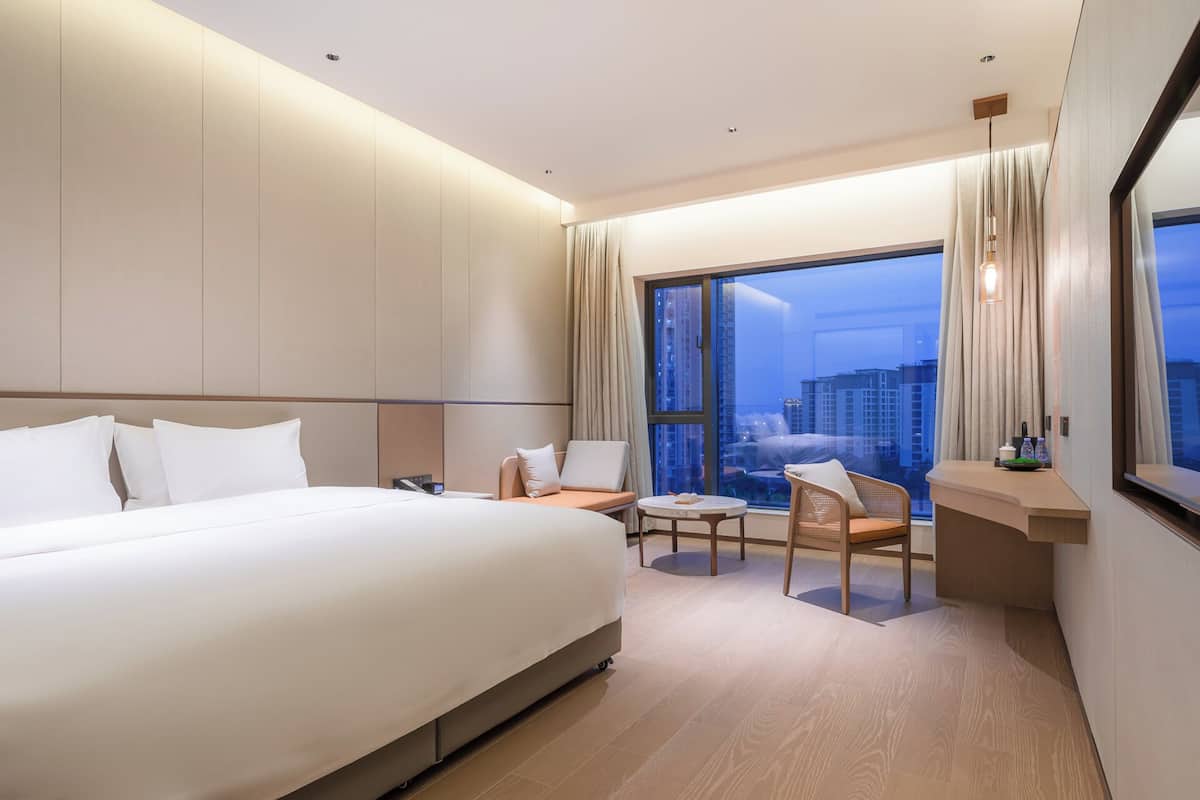 A modern hotel room with a large bed, a seating area with a table, and a view of the cityscape through large windows.