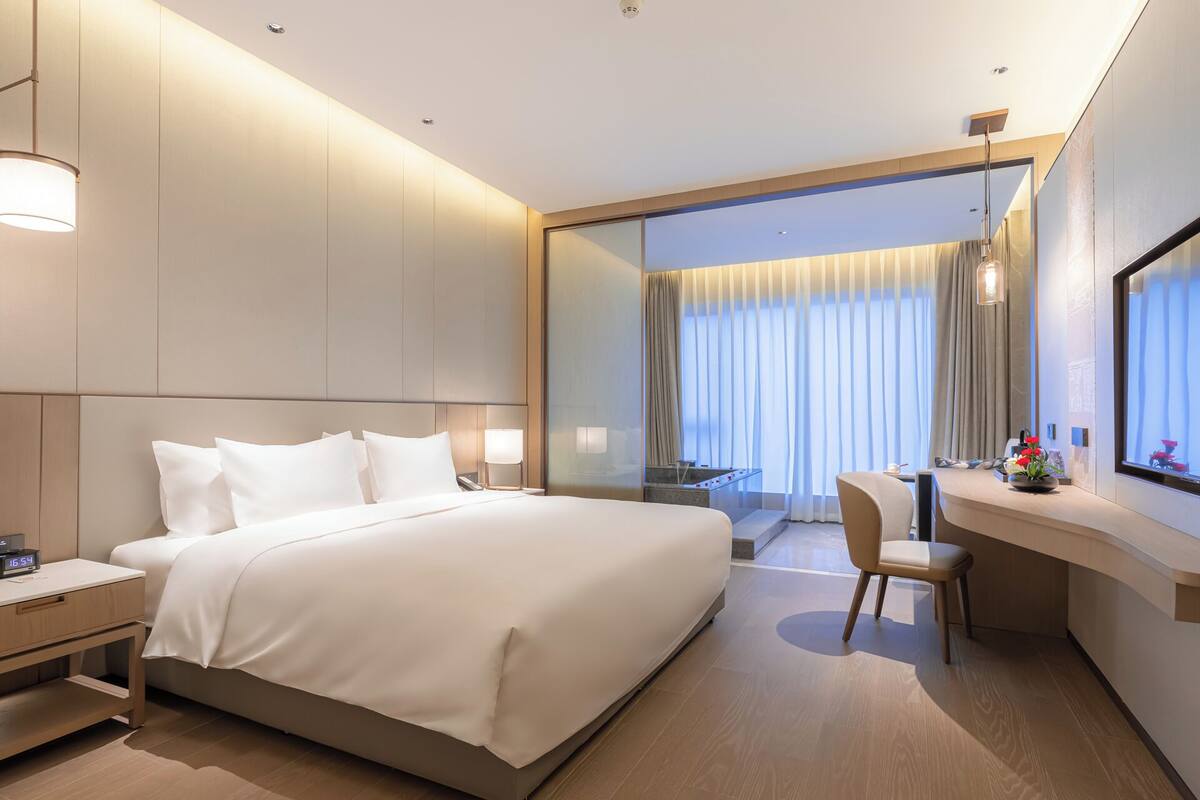 A modern hotel room with a large bed, bedside tables, a desk with a chair, a large mirror, and a view of the outdoors through a glass door.