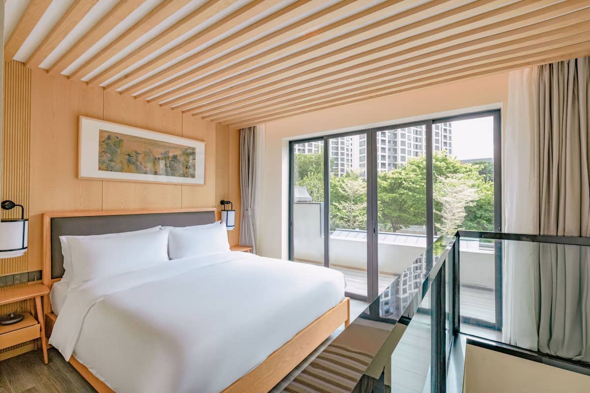 A modern hotel room with a large bed, a painting on the wall, and a balcony with a city view.